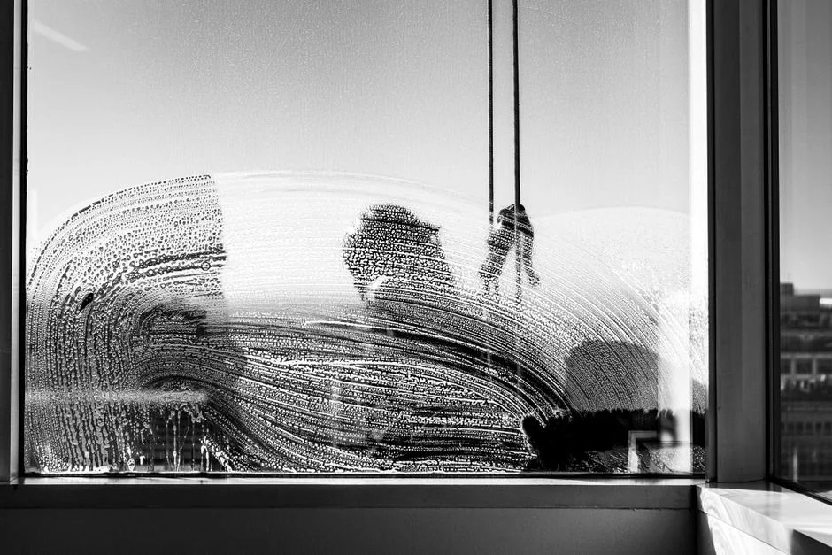 Black and white from below of man steeplejack in protective helmet with professional equipment cleaning glass wall of modern tower